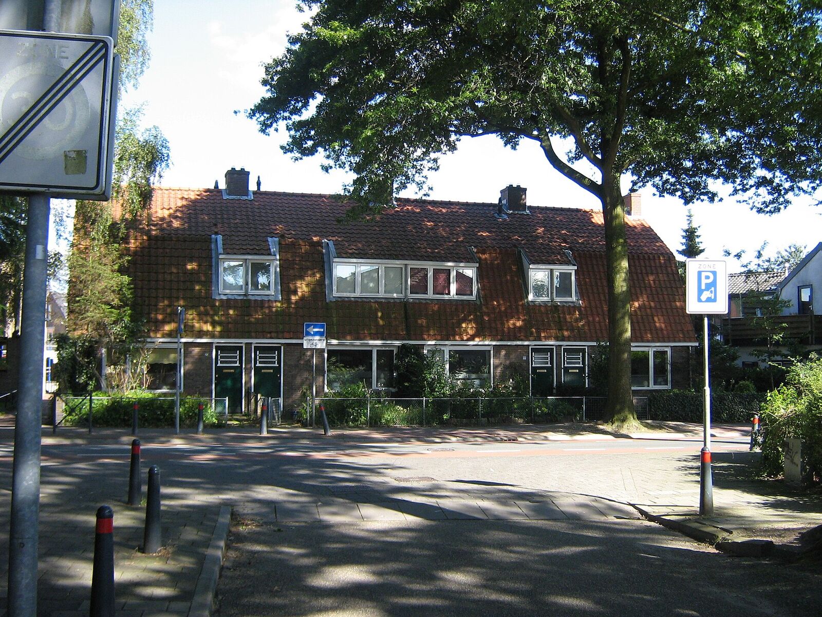 A row of brick houses with red-tiled roofs and white window frames behind a hedge and a large tree, on the Bosdrift in Hilversum