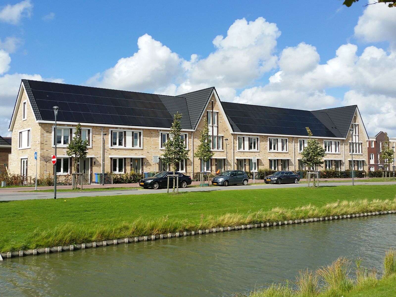 A terrace of new yellow-brick houses with solar panels built into the dark roof, seen across a canal and a strip of grass